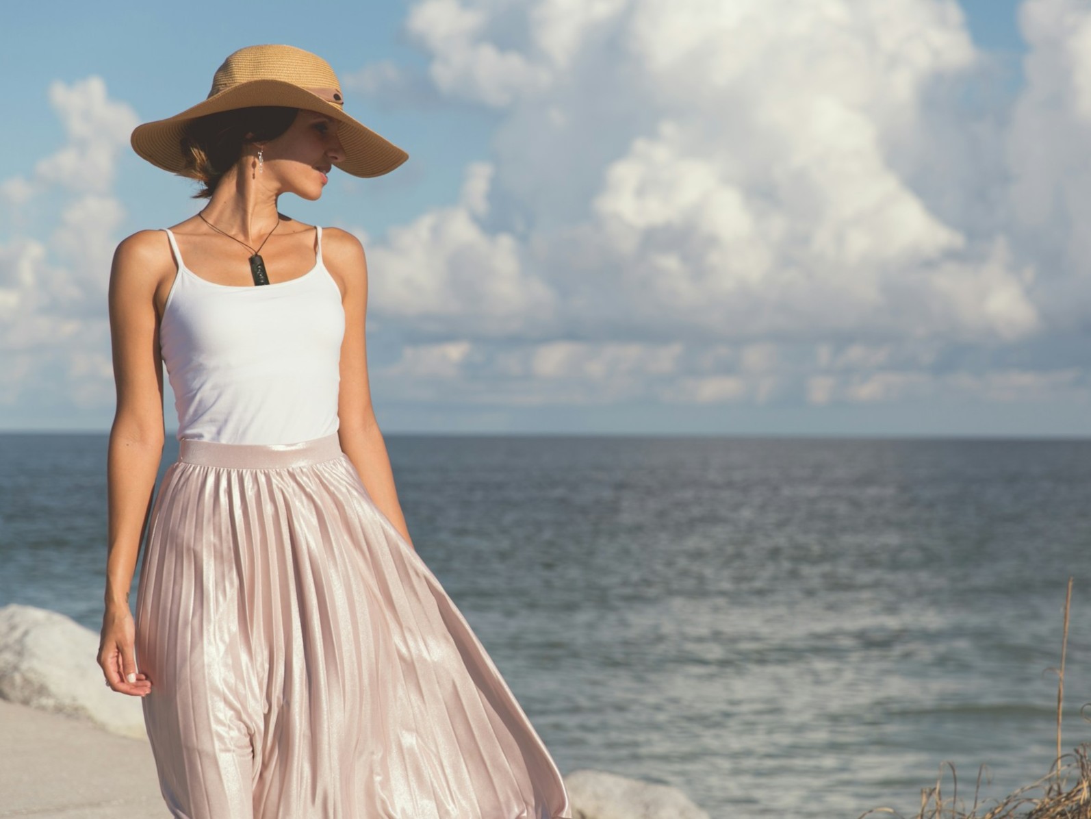 Woman in a straw hat and pink pleated skirt standing by the ocean under a cloudy sky.